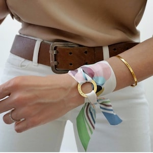 May include: A silk scarf tied through a gold-toned metal ring, featuring a multi-colored abstract pattern of pink, blue, green, and black shapes. The scarf is wrapped around a wrist, complemented by a thin gold-toned metal bangle and a ring.