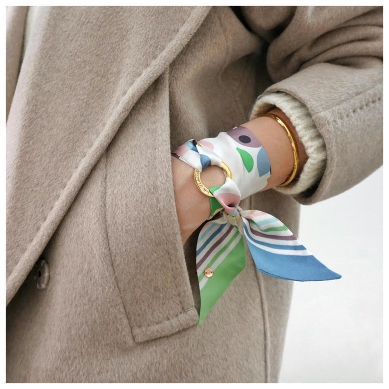 May include: A silk scarf tied around a gold-toned metal ring, featuring a white base with abstract shapes in pink, purple, blue, and green, alongside a section with thin horizontal stripes in matching colors, worn over a beige wool coat.