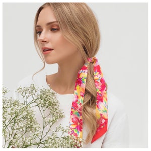 May include: A woman with long blonde hair is wearing a white shirt and a floral patterned hair scarf. The scarf is tied in a bow at the back of her head. The scarf is pink, red, yellow, and green.