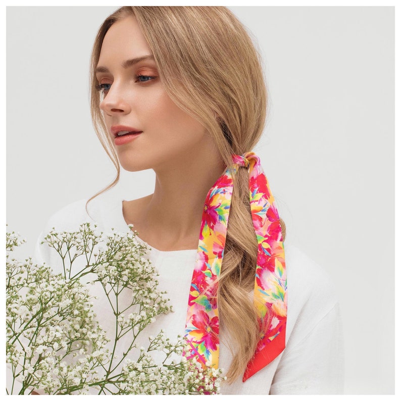 May include: A woman with long blonde hair is wearing a white shirt and a floral patterned hair scarf. The scarf is tied in a bow at the back of her head. The scarf is pink, red, yellow, and green.