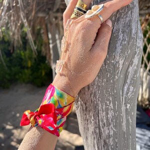 May include: A red and blue patterned silk scarf tied around a wrist. The scarf has a floral design with yellow, green, and pink accents. The scarf is tied in a bow.