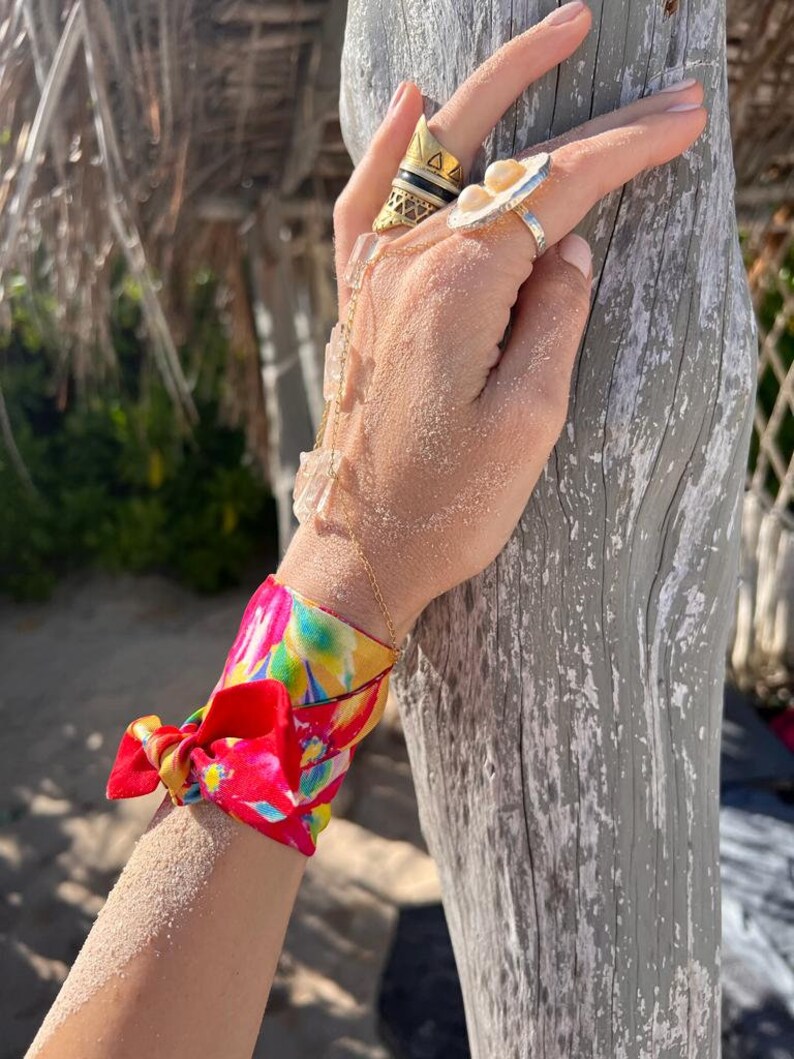 May include: A red and blue patterned silk scarf tied around a wrist. The scarf has a floral design with yellow, green, and pink accents. The scarf is tied in a bow.