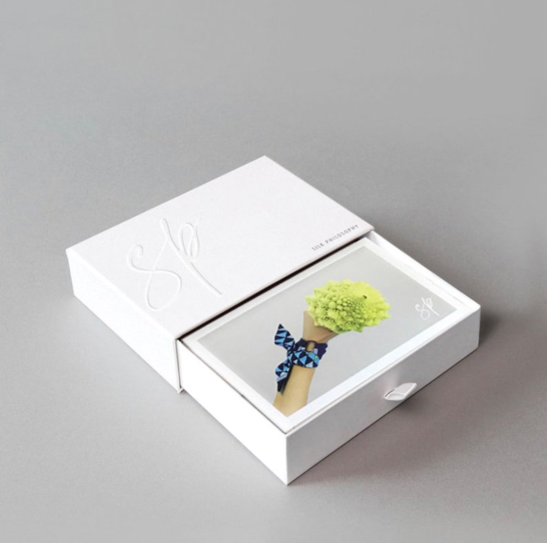 May include: White box with a drawer that slides out. The box has the word "She" embossed on the top and the words "Silk Philosophy" printed on the side. The drawer has a white background and a photo of a hand holding a green flower with a blue and white patterned ribbon tied around the wrist.
