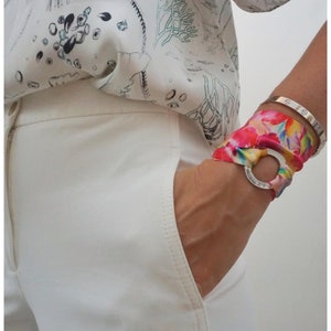 May include: White pants with a pocket and a colorful patterned scarf tied around the wrist with a silver ring.
