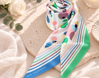 May include: A white silk scarf featuring a colorful pattern of scattered circles in pink, blue, green, and yellow, with striped borders in blue and green. The text SILK PHILOSOPHY is printed in white near the pointed end of the scarf.