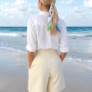 May include: A white long-sleeved button-down shirt with rolled cuffs, paired with cream-colored tailored shorts featuring back pockets. A patterned hair scarf with blue, green, and purple geometric shapes and stripes is tied around a ponytail.