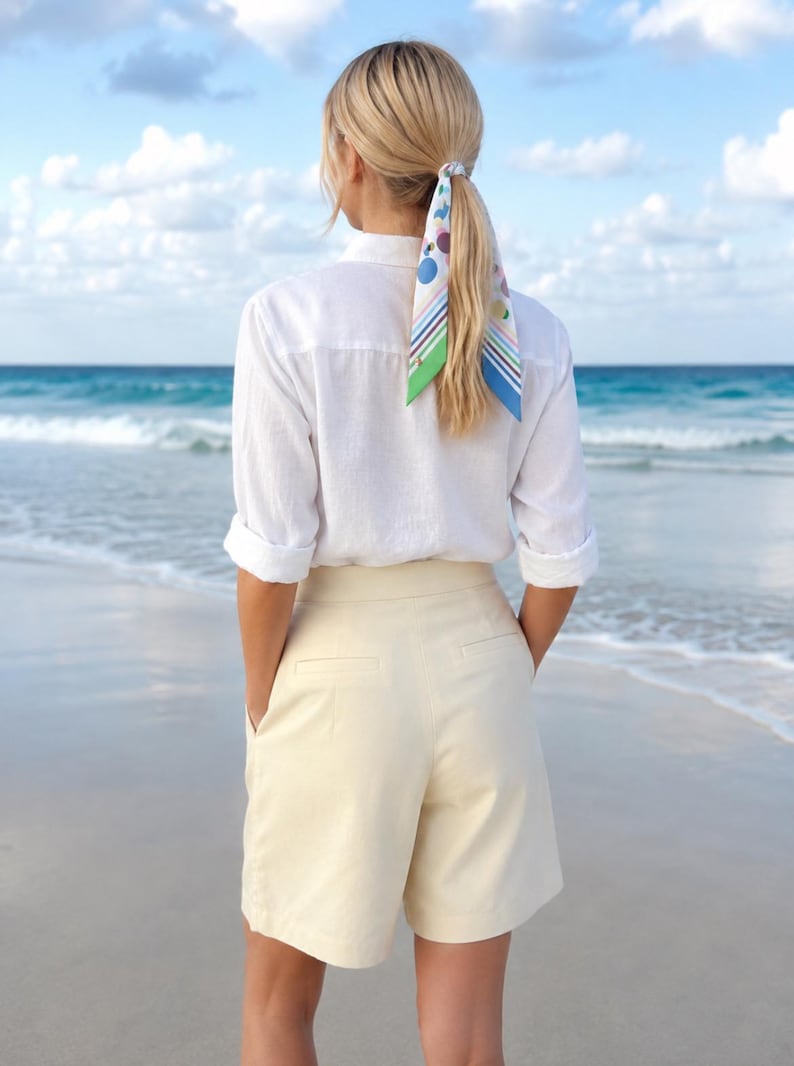 May include: A white long-sleeved button-down shirt with rolled cuffs, paired with cream-colored tailored shorts featuring back pockets. A patterned hair scarf with blue, green, and purple geometric shapes and stripes is tied around a ponytail.