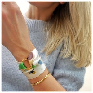 May include: A gold-toned metal ring bracelet featuring a white silk scarf threaded through the center. The scarf displays a multi-colored abstract pattern with shapes in green, blue, brown, and yellow. A thin, plain gold-toned metal bangle is worn below.