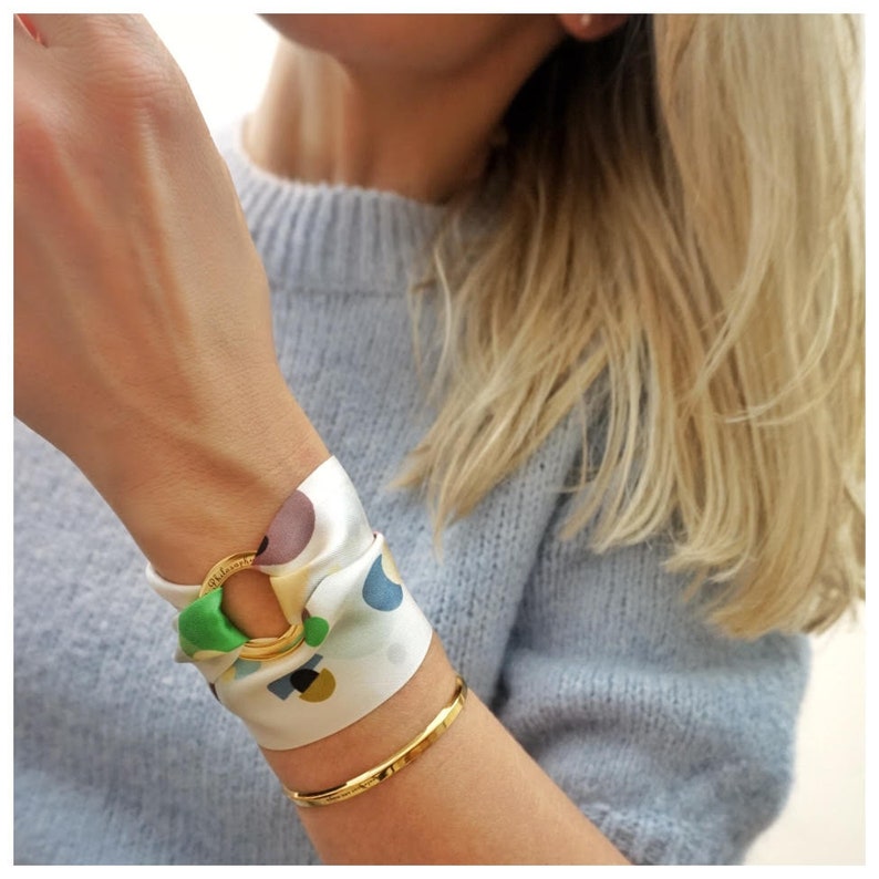 May include: A gold-toned metal ring bracelet featuring a white silk scarf threaded through the center. The scarf displays a multi-colored abstract pattern with shapes in green, blue, brown, and yellow. A thin, plain gold-toned metal bangle is worn below.