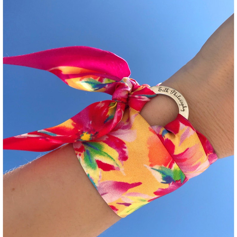 May include: A colorful floral patterned silk scarf tied around a wrist with a silver charm that says "Silk Philosophy".