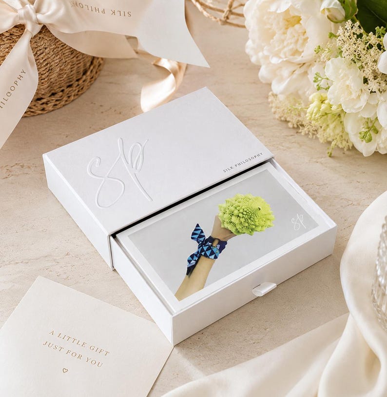 May include: A white rectangular sliding gift box with the embossed letters SP and the text SILK PHILOSOPHY. Inside, a card displays a graphic of a wrist wearing a blue patterned silk ribbon and holding a bright green romanesco broccoli.