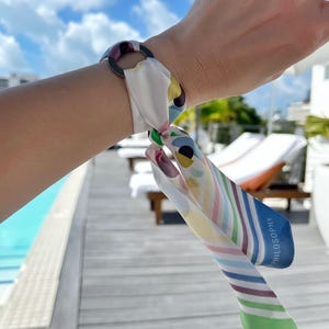 May include: A mink silk scarf tied through a circular ring, featuring a multicolored pattern of abstract shapes, dots, and diagonal stripes in shades of blue, green, yellow, and pink, with the word SILK PHILOSOPHY printed on the blue-bordered edge.