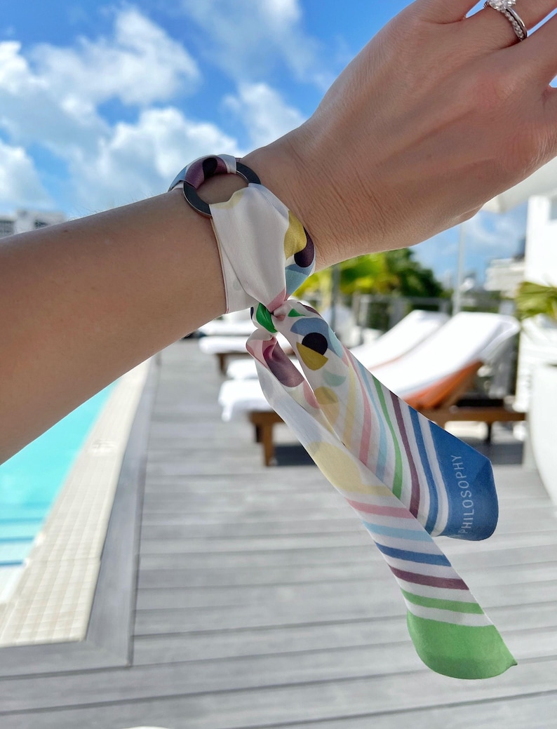 May include: A mink silk scarf tied through a circular ring, featuring a multicolored pattern of abstract shapes, dots, and diagonal stripes in shades of blue, green, yellow, and pink, with the word SILK PHILOSOPHY printed on the blue-bordered edge.
