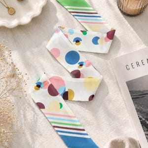 May include: A long, narrow silk scarf featuring a white base with a colorful pattern of scattered circles in shades of blue, pink, yellow, and maroon, complemented by striped ends in green, blue, pink, and yellow, with pointed, angled edges.