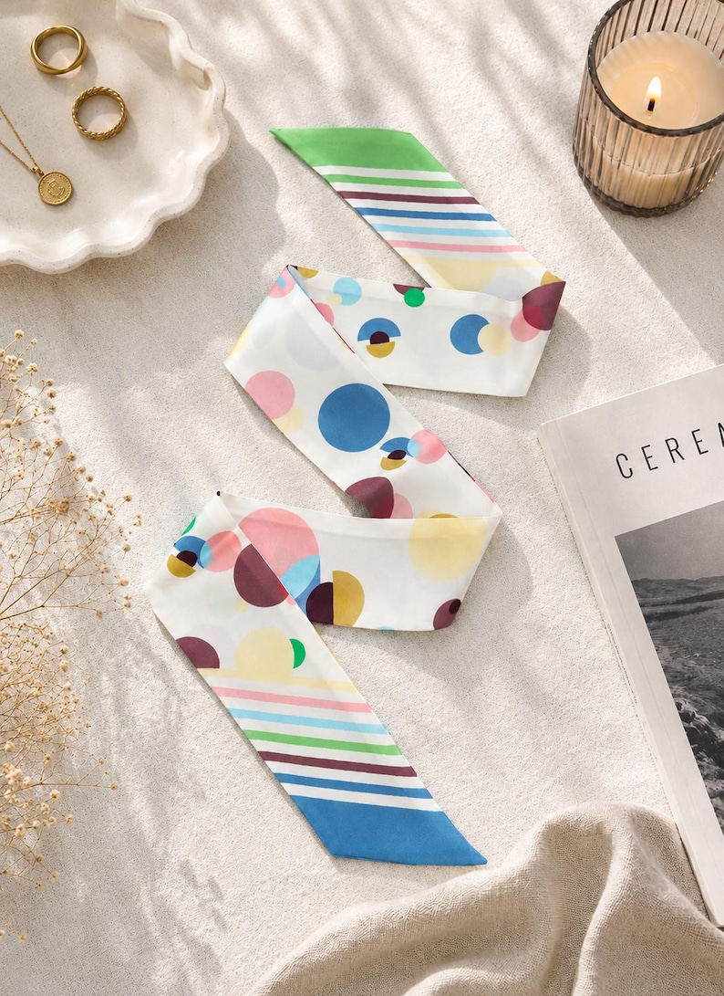 May include: A long, narrow silk scarf featuring a white base with a colorful pattern of scattered circles in shades of blue, pink, yellow, and maroon, complemented by striped ends in green, blue, pink, and yellow, with pointed, angled edges.