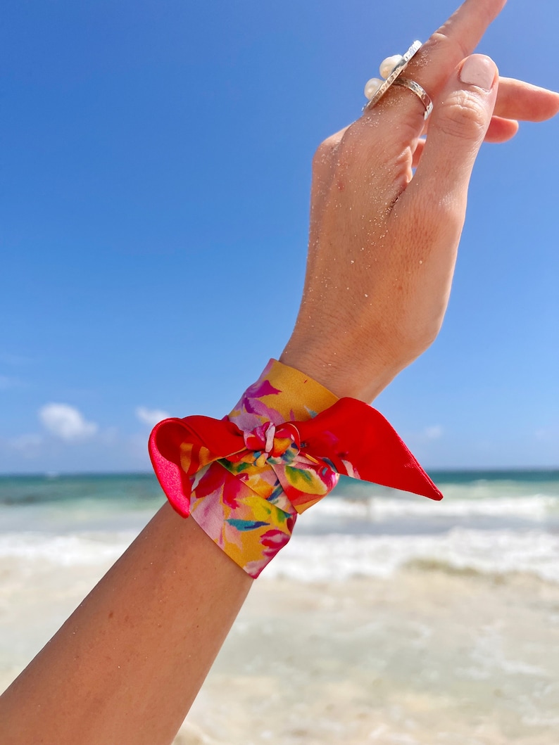 May include: A red and yellow floral patterned silk scarf tied around a wrist as a bracelet. The scarf is tied in a knot with the ends of the scarf hanging down.
