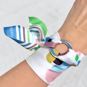 May include: A white silk bracelet featuring a metallic ring and a colorful pattern of abstract shapes in blue, green, yellow, and pink. The fabric is tied into a bow, and the text "SILK PHILOSOPHY" is printed on the ring and the blue fabric edge.