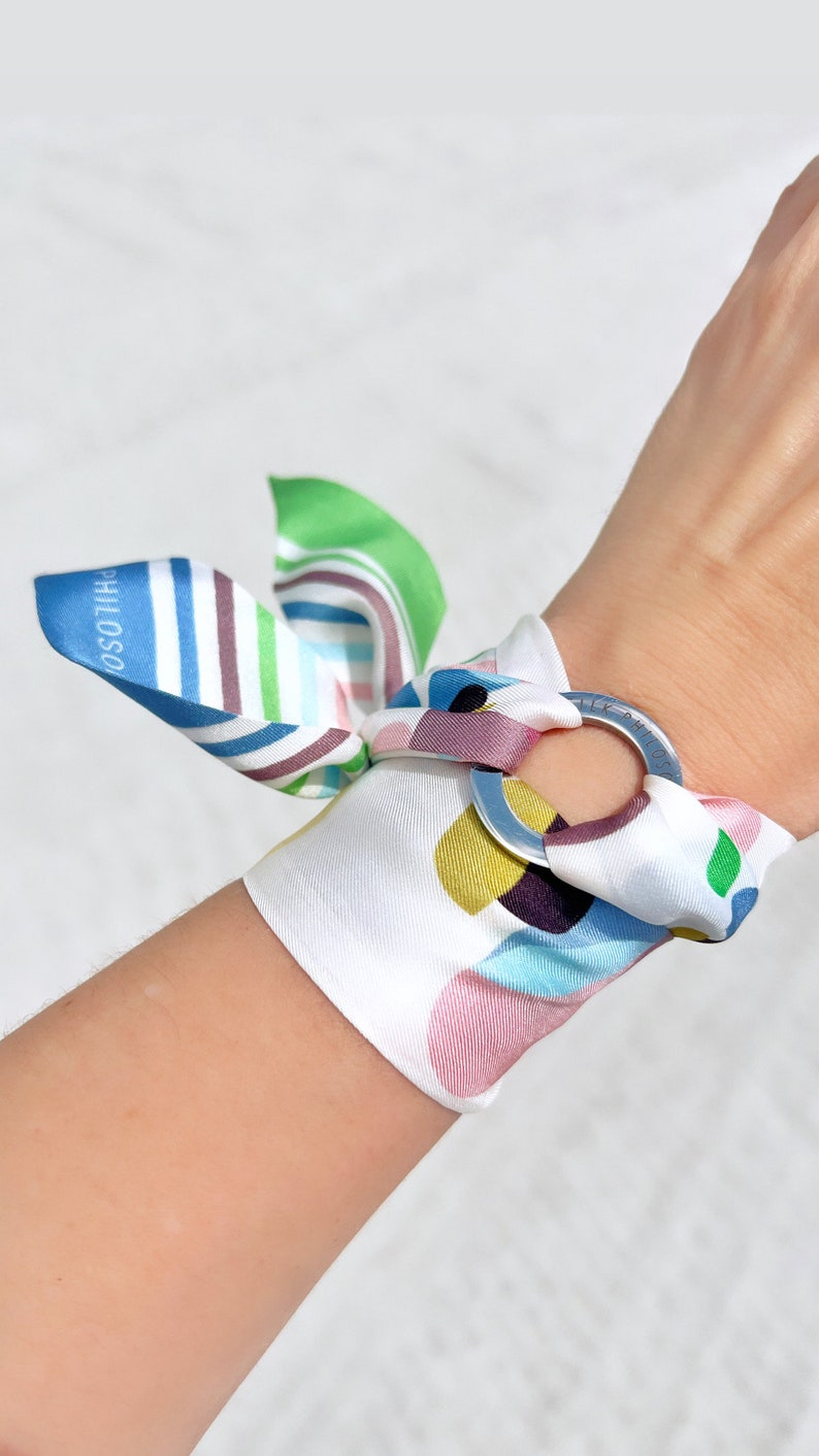 May include: A white silk bracelet featuring a metallic ring and a colorful pattern of abstract shapes in blue, green, yellow, and pink. The fabric is tied into a bow, and the text "SILK PHILOSOPHY" is printed on the ring and the blue fabric edge.