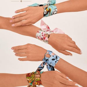 May include: Four silk wrist scarves tied through circular metal rings. The scarves feature various patterns including abstract blue and yellow shapes, pink floral prints, light blue floral designs, and multicolored autumn leaf motifs.