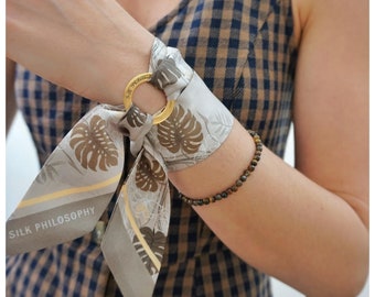 May include: A light gray silk scarf featuring a brown monstera leaf pattern and a gold-toned metal ring engraved with the text Silk Philosophy. The scarf is tied through the ring, creating a decorative wrist accessory alongside a beaded bracelet.