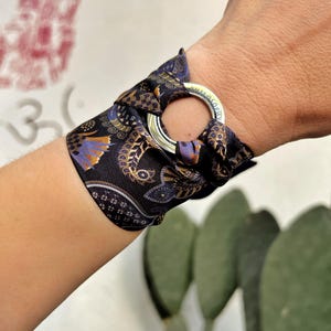 Può includere: Un braccialetto in seta nera e oro con un anello d'argento. Il braccialetto presenta un motivo a paisley con accenti viola e oro. L'anello d'argento ha incisa la parola "PHILOSOPHY". Il braccialetto è avvolto attorno a un polso.