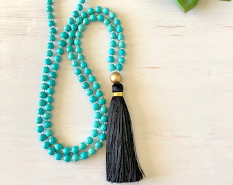 May include: A turquoise blue beaded necklace with a black tassel and gold accent. The necklace is on a white wooden surface.