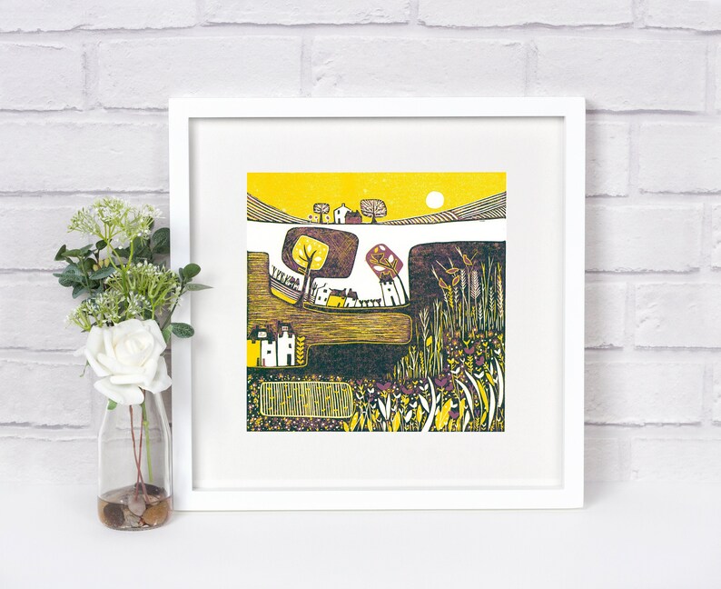 May include: A colorful linocut print of a rural landscape with houses, trees, and fields. The print is framed in white and has a yellow background.