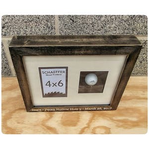 Puede incluir: Un marco de madera oscura con un acabado envejecido que contiene una pelota de golf y un espacio de 4x6 pulgadas para una foto. El texto en el marco dice "Eagle - Pecan Hollow Hole 9 - March 26, 2017."