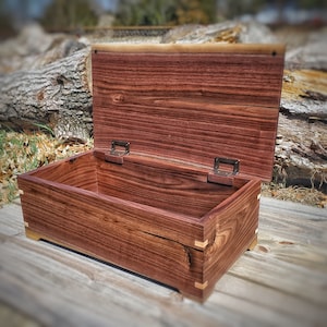 May include: A wooden box with a hinged lid, crafted from dark wood with a rich grain pattern. The box has brass hinges and a natural wood finish. The box is open, revealing the empty interior.