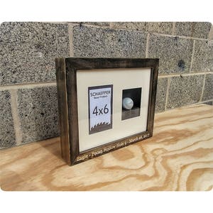 Puede incluir: Un marco de exhibici&oacute;n con una pelota de golf y un espacio para fotos de 4x6. El marco es de color marr&oacute;n oscuro con un acabado envejecido. El texto en el marco dice "Eagle - Pecan Hollow Hole 9 - March 26, 2017." El marco est&aacute; sobre una superficie de madera.