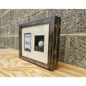 Puede incluir: Una exhibici&oacute;n enmarcada con un marco de madera te&ntilde;ida oscura. La exhibici&oacute;n presenta una abertura para fotos de 4x6 y un &aacute;rea empotrada que contiene una pelota de golf. El texto "SCHAEFFER Wood Products" y "Eagle - Pecan Hollow Hole 9 - March 26, 2017" son visibles.