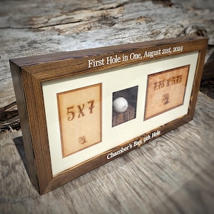 May include: A wooden framed display featuring a golf ball and two wooden plaques. The plaques are labeled "5X7" and "7.75 X 5.75". The frame includes the text "First Hole in One, August 21st, 2024" and "Chamber's Bay, 9th Hole".