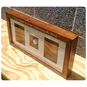 May include: A wooden frame with a glass front displays a golf ball. The frame is engraved with the text "Jim's Hole In One / February 8, 2014 Whirlwind Golf Club / Devils Claw / 14th Hole".
