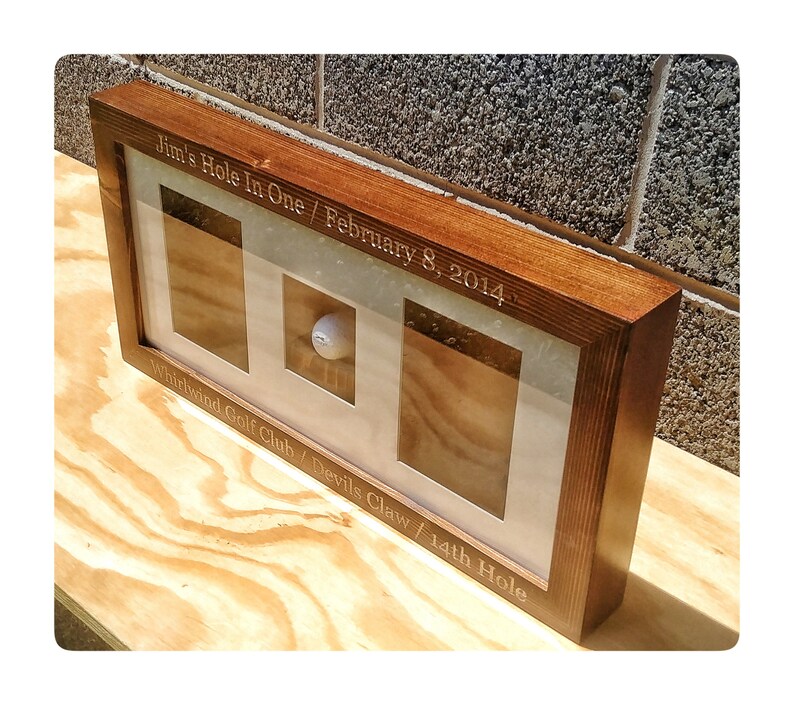 May include: A wooden frame with a glass front displays a golf ball. The frame is engraved with the text "Jim's Hole In One / February 8, 2014 Whirlwind Golf Club / Devils Claw / 14th Hole".