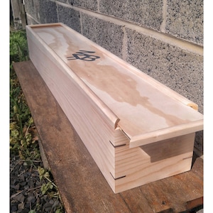 May include: A long, rectangular wooden box with a hinged lid. The box is made of light-colored wood and has a natural finish. The lid has a simple design with a raised edge and a small, black inscription on the top.