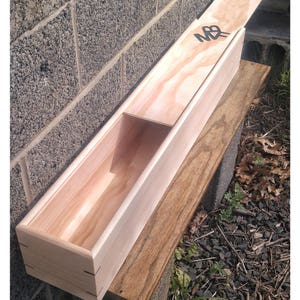 May include: A wooden box with a hinged lid, the lid is open and the box is resting on a wooden surface. The box is made of light wood and has a natural finish. The lid has a black letter "M" and a black letter "B" on it.