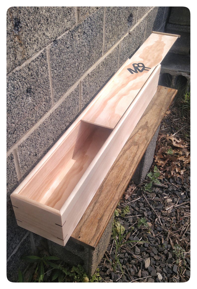 May include: A wooden box with a hinged lid, the lid is open and the box is resting on a wooden surface. The box is made of light wood and has a natural finish. The lid has a black letter "M" and a black letter "B" on it.