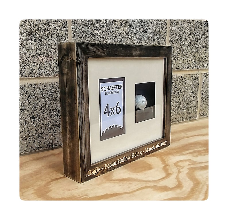 Puede incluir: Un marco de madera r&uacute;stico con un acabado oscuro, con un espacio para fotos de 4x6 y un compartimento para una pelota de golf. El marco incluye el texto "SCHAEFFER Wood Products" y "Eagle - Pecan Hollow Hole 9 - March 26, 2017".