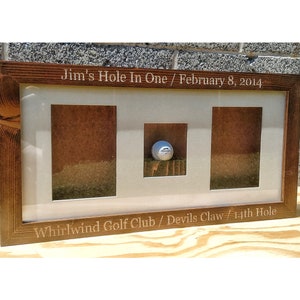 May include: A wooden frame with a golf ball inside. The frame has an inscription that reads "Jim's Hole In One / February 8, 2014 Whirlwind Golf Club / Devils Claw 14th Hole".
