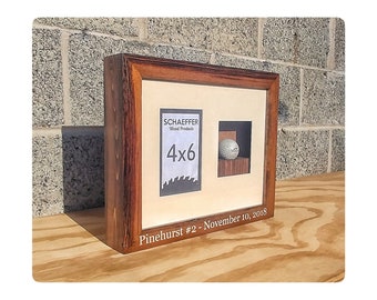 May include: A wooden picture frame with a golf ball inside. The frame is engraved with "Pinehurst #2 - November 10, 2018". The frame also has a label that says "Schaeffer Wood Products 4x6".