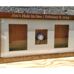 May include: A wooden frame with a golf ball displayed in the center. The frame is engraved with the text "Jim's Hole In One / February 8, 2014 Whirlwind Golf Club / Devils Claw / 14th Hole".