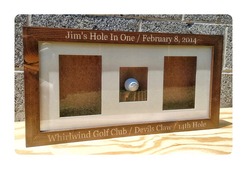 May include: A wooden frame with a golf ball displayed in the center. The frame is engraved with the text "Jim's Hole In One / February 8, 2014 Whirlwind Golf Club / Devils Claw / 14th Hole".