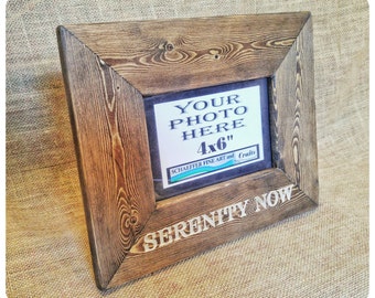 May include: A rustic wooden picture frame with a 10.2 x 15.2 cm opening. The frame is stained a dark brown colour and has a distressed finish. The text "YOUR PHOTO HERE" is printed on the frame, along with the text "SERENITY NOW".