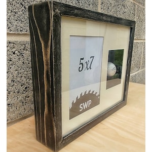 May include: A black wood picture frame with a white mat and a 5x7 inch photo opening. The frame has a saw blade design with the text "SWP" and a small, clear, square opening for a golf ball.