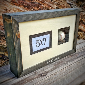 May include: A dark gray wood picture frame with a white mat and a 5x7 inch photo opening. The frame has a small opening for a baseball, which is included in the frame. The frame is engraved with the date "July 16, 2023".