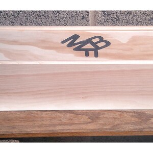 May include: A long, rectangular wooden box with a light wood finish. The box has a hinged lid and a black logo on the top that says "MB".