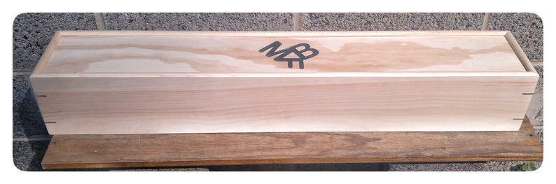 May include: A long, rectangular wooden box with a light wood finish. The box has a hinged lid and a black logo on the top that says "MB".