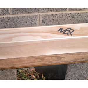 May include: A long, wooden box with a hinged lid. The box is made of light wood and has a small compartment on the left side. The box is labelled with the initials "MB" in black lettering.