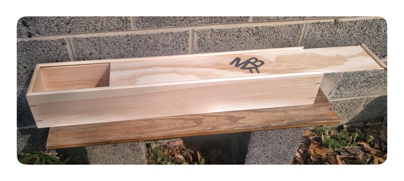May include: A long, wooden box with a hinged lid. The box is made of light wood and has a small compartment on the left side. The box is labelled with the initials "MB" in black lettering.
