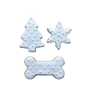 May include: Three white sugar cookies decorated with white dots. The cookies are shaped like a Christmas tree, a snowflake, and a bone.